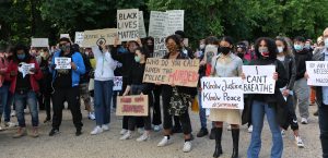 Man sieht mehrere Menschen mit Plakaten eine Straße entlang laufen; auf den Plakaten stehen Botschaften gegen Rassismus und Diskriminierung