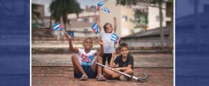 Drei Kinder sitzen auf dem Boden und halten kubanische Flaggen in den Händen. Hinter ihnen sieht man ein paar Häuser und der rechte Junge hält einen Roller in der Hand.