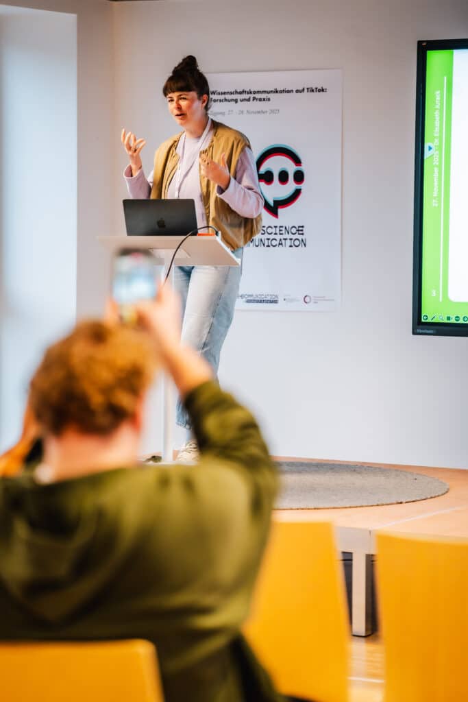 Eine Frau steht auf der Bühne. Vor ihr, nur verschwommen zu sehen, filmt sie jemand mit dem Smartphone.